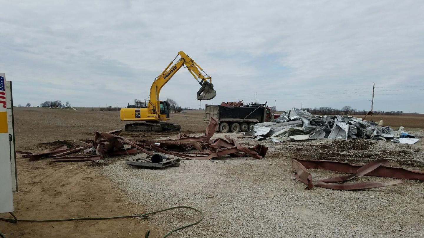 Picture of the metal farm building completely torn down and seperated, being loaded into dump trucks.